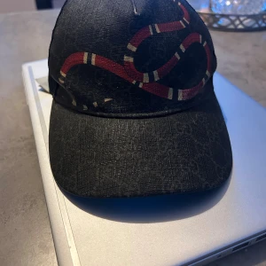 Gucci svart keps med ormprint - Svart keps från Gucci med mesh på sidorna och bak, samt ett coolt rött och vitt ormprint på framsidan. Kepsen har diskret GG-monogram över hela tyget och justerbar rem bak. Perfekt för dig som vill sticka ut med designeraccessoar.