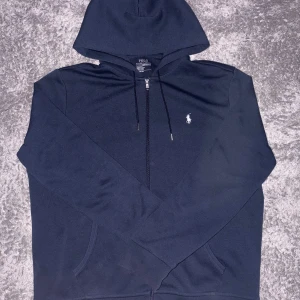 Marinblå hoodie från Polo Ralph Lauren - Snygg marinblå hoodie från Polo Ralph Lauren med klassisk vit logga på bröstet. Tröjan har huva med snörning, hel dragkedja och två fickor framtill. Tillverkad i mjuk bomullsmix som är skön att ha på sig. Perfekt för chill dagar och streetwear vibes.