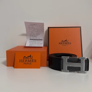 Svart H bälte med H-spänne - Exklusivt svart bälte från H med klassiskt H-spänne i silverfärgad metall. Bältet är tillverkat i präglat skinn med en stilren och modern look. Levereras med originalförpackning och Hermès-kort. Perfekt accessoar för dig som gillar lyxiga detaljer.