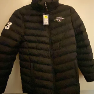Svart pufferjacka Vinson Original - Svart pufferjacka från Vinson Original med vit logga på bröstet och stort tryck '23' på ena ärmen. Jackan har dragkedja framtill och är vadderad för extra värme. Perfekt för kalla dagar och har en sportig vibe.