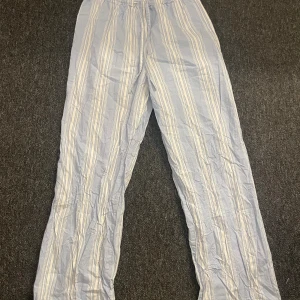 Randiga bomullsbyxor från H&M - Ljusblå och vita randiga byxor från H&M i storlek S. Byxorna har elastisk midja med snörning och är tillverkade i 100% bomull. De har en avslappnad passform och raka ben, perfekta för chill dagar eller som mjukisbyxor.