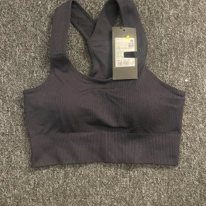 Svart ribbad sporttopp från Tridri - Svart ribbad sporttopp från Tridri med bred resår under bysten och brottarrygg. Toppen är sömlös och har en tight passform som ger bra stöd vid träning. Materialet är stretchigt och består av 92% polyamid och 8% elastan.