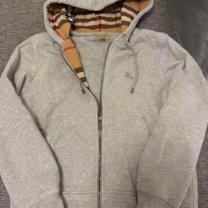 Grå hoodie från Burberry - Säljer en grå hoodiejacka från Burberry med klassiskt rutigt foder i beige, svart, rött och vitt. Hoodien har dragkedja, snörning i huvan och ribbade muddar. Perfekt för dig som gillar stilrena detaljer och ikoniskt mönster.