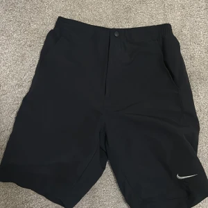 Svarta Nike shorts med fickor - Svarta shorts från Nike med elastisk midja och knapp framtill. Klassisk design med två sidofickor och en bakficka. Perfekta för träning eller chill dagar. Liten Nike-logga i vitt på ena benet. Lätta och bekväma att bära.