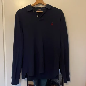 Marinblå långärmad pikétröja Polo Ralph Lauren - Snygg marinblå långärmad pikétröja från Polo Ralph Lauren med klassisk krage och två vita knappar. Tröjan har det ikoniska röda broderade Polo-logot på bröstet och är gjord i mjuk bomull. Perfekt för dig som gillar stilren och tidlös design. Priset är ej hugget i sten! Kom gärna med frågor.