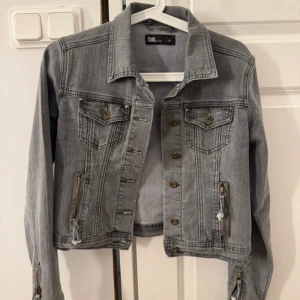 Grå jeansjacka med dragkedjor från Flash - Snygg grå jeansjacka från Flash med coola detaljer som dragkedjor vid ärmslut och fickor samt nitar på ryggen. Klassisk krage, knappar framtill och två bröstfickor. Perfekt för dig som gillar en edgy stil.