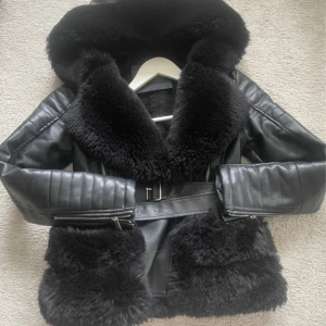 Svart jacka med bälte och dragkedja - Snygg svart pälsjacka med bred krage och fluffiga detaljer på krage och nederkant. Jackan har quiltade skinnärmar, bälte i midjan och dragkedjor vid fickorna. Perfekt för dig som vill ha en statementjacka med lyxig vibe och cool siluett. Storlek S. Men den är ganska tight. Jag brukar ha storlek Xs-s  Nypris: 2400kr