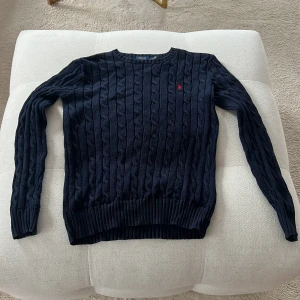 Mörkblå kabelstickad tröja Polo Ralph Lauren - Kabelstickad mörkblå tröja från Polo Ralph Lauren med klassisk röd broderad logga på bröstet. Tröjan har rund halsringning, långa ärmar och ribbade muddar. Perfekt för lager-på-lager och snygg till jeans