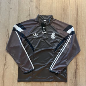 Adidas Real Madrid half zip tröja - Snygg grå och svart långärmad Adidas-tröja med Real Madrid-logga på bröstet. Tröjan har half zip, vita detaljer och klassiska Adidas-ränder längs ärmarna. Perfekt för fotboll eller chill. Tillverkad i polyester för en sportig känsla.