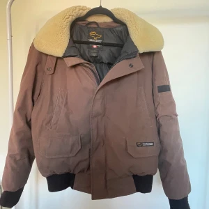 Brun Canada goose jacka GE BUD  - Säljer en brun bomberjacka från Canada Goose med stor beige fårkrage och svart quiltat innerfoder. Jackan har klassisk patch på ärmen, dragkedja och muddar vid ärmslut och midja. Perfekt för kalla dagar och riktigt snygg streetstil.