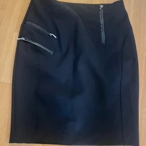 Svart kjol med dragkedjor - Snygg svart pennkjol med tre coola dragkedjor framtill som ger en edgy vibe. Kjolen är figurnära och har en stilren design utan mönster. Perfekt för dig som gillar minimalistisk stil med en twist.