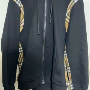 Burberry svart hoodie med rutiga detaljer - Svart hoodie från Burberry med klassiska beige-rutiga paneler längs ärmarna. Tröjan har huva med snörning, dragkedja framtill och känguruficka. Tillverkad i mjuk bomull och har en clean, oversized vibe som sticker ut med de ikoniska rutorna.