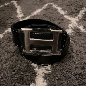 H-bälte  - Snyggt bälte i  svart skinn med ikoniskt,H-spänne i silverfärgad metall. Bältet har stilrena, kontrastsömmar och klassisk design som lyfter, vilken outfit som helst. Strl 110cm