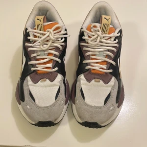 Puma RS X i flera färger - Säljer sonens Puma RS-X running system unisex sneakers med cool retrostil. Skorna har vit mesh, grå mockadetaljer, svarta och lila paneler samt orange och beige inslag. Tjock sula och snörning framtill. Perfekta för dig som gillar statement sneakers med sportig vibe. Skorna använts sparsamt få gånger.