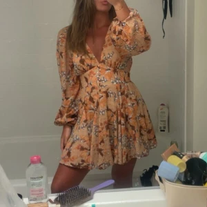 Blommig kortklänning med volang - Superfin kortklänning med blommigt mönster i beige och orange toner. Klänningen har v-ringad hals, volang i kjolen och långa puffärmar. Perfekt för sommarens alla tillfällen, med omlott-effekt och skön passform.