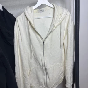 Vit hoodie med dragkedja Studio Total - Snygg vit hoodie från Studio Total i storlek M. Tröjan har huva med snörning, praktiska fickor och hel dragkedja framtill. Tillverkad i mjuk bomullsmix som är skön mot huden. Perfekt för chill dagar och enkel att matcha med jeans eller joggers.
