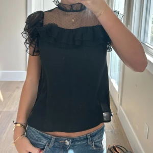 Svart blus med volang och mesh - Säljer en svart blus med transparent mesh upptill och små prickar. Blusen har volangdetaljer vid axlar och hals, samt en hög krage med rynk. Använd en gång. Obs, smuts på spegeln inte tröjan!