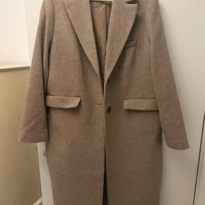 Beige kappa med klassisk krage - Snygg beige kappa med klassisk krage och knäppning framtill. Jackan har två fickor med lock och en slits bak för extra rörelsefrihet. Långa ärmar med knappar vid ärmslut och stilren, rak passform. Perfekt för dig som gillar minimalistisk och clean stil.