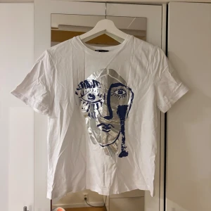 Vit t-shirt med silvertryck från Zara - Vit t-shirt från Zara i bomull med korta ärmar och ett stort konstnärligt ansikte tryckt i silver och mörkblått framtill. Skön passform och cool design som sticker ut. Perfekt för dig som gillar unika prints och streetstyle.
