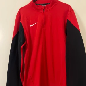 Röd Nike Dri-Fit half zip tröja - Röd långärmad Nike Dri-Fit tröja med svarta ärmar och vit logga på bröstet. Tröjan har en half zip-dragkedja vid halsen och är tillverkad i ett lätt och funktionellt material som passar perfekt för träning eller sport.
