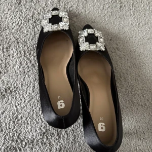 Svarta pumps med pärldetalj, strl 38 - Eleganta svarta pumps med spetsig tå och låg klack. Skorna är tillverkade i satinliknande material och har en stor dekorativ pärlbesatt brosch framtill som ger extra lyxig känsla. Perfekt för dig som vill sticka ut med stilrena detaljer.