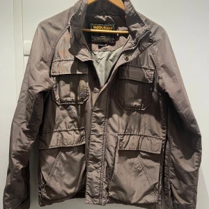 Brun vindjacka från Woolrich, stl S - Säljer en brun vindjacka från Woolrich med flera stora fickor framtill och dragkedja. Jackan har hög krage och klassisk outdoor-look, perfekt för dig som gillar funktionella plagg med stil. Märke och detaljer syns tydligt på insidan.
