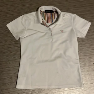 Vit piké från Gant med ränder vid nack kragen  - Vit pikétröja från Gant med klassisk krage och korta ärmar. Insidan av kragen har färgglada ränder som ger en snygg kontrast. Broderad Gant-logga med sköld på bröstet. Perfekt för en clean och sportig look.