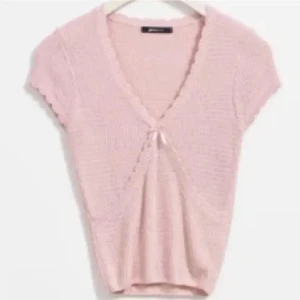 Ljusrosa stickad bolerotopp med rosett - Supersöt ljusrosa stickad bolerotopp med korta ärmar och v-ringning. Toppen har en liten rosett framtill och ribbad kant vid midjan. Perfekt för dig som gillar mjuka pasteller och romantisk stil. Liten i storleken.