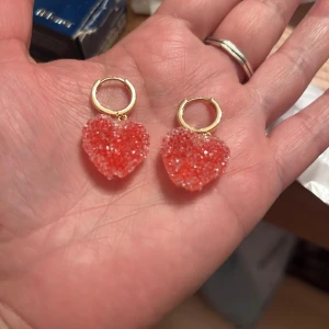 Guldiga örhängen med röda hjärta - Säljer ett par guldiga hoops med en glittrig röda hjärtberlock. Hjärtat är gjort av små pärlliknande detaljer som ger en cool textur och extra bling. Perfekt accessoar för dig som vill sticka ut med något sött och trendigt.