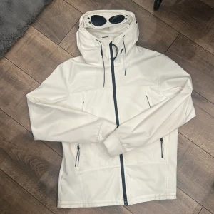 Vit Goggle-jacka från C.P. Company - Säljer en clean och stilren vit Goggle-jacka från C.P. Company med ikoniska glasögon i huvan. Jackan har hel dragkedja, två sidofickor med dragkedja och justerbar huva med snörning. Tillverkad i stretchigt soft shell-material med återvunnen nylon.