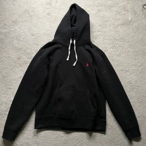 Svart hoodie från Polo Ralph Lauren - Svart hoodie från Polo Ralph Lauren med klassisk röd broderad logga på bröstet. Tröjan har vit snörning i huvan, känguruficka och är tillverkad i mjuk bomull. Perfekt för en chill och stilren look. Nyprics ca 1800kr