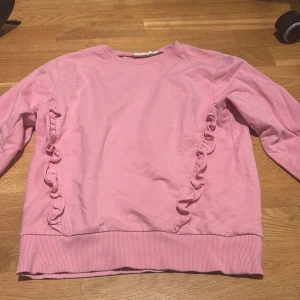 Rosa sweatshirt med volangdetaljer - Säljer en söt rosa sweatshirt med volangdetaljer längs sidorna. Tröjan har rund halsringning, långa ärmar och ribbade muddar vid ärmslut och nederkant. Perfekt för dig som gillar en feminin och avslappnad stil.
