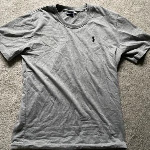 Grå t-shirt från Polo Ralph Lauren - Snygg grå t-shirt från Polo Ralph Lauren med klassisk rund halsringning och broderad svart logga på bröstet. Tillverkad i mjuk bomull och har en normal passform. Perfekt basic-plagg med stilren look.