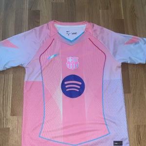 FC Barcelona rosa matchtröja Nike - Snygg FC Barcelona fotbollströja från Nike i rosa och ljusblå toner. Tröjan har korta ärmar, V-ringning och tryckta detaljer som klubbmärke och Spotify-logga. Tillverkad i lätt och ventilerande funktionsmaterial, perfekt för träning eller match.