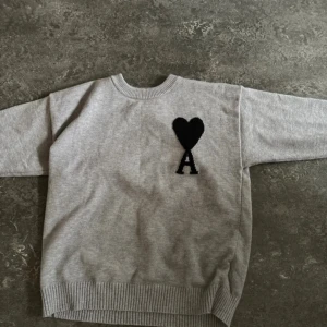 Grå stickad tröja från Ami Paris - Säljer en grå stickad tröja från Ami Paris med svart hjärta och bokstaven A broderat på bröstet. Tröjan har rund halsringning, ribbade muddar och är långärmad. Perfekt för dig som gillar stilrena och trendiga plagg.
