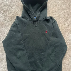 Svart hoodie från Polo Ralph Lauren - Svart hoodie från Polo Ralph Lauren med klassisk röd broderad logga på bröstet. Tröjan har huva, känguruficka och långa ärmar. Tillverkad i mjuk bomull och har en normal passform. Det står XL på insidan men den passar XS/S.