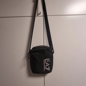 Svart axelväska från Emporio Armani - Snygg svart axelväska från Emporio Armani EA7 med justerbar rem och dragkedja. Väskan har en stilren design med logga framtill och är tillverkad i slitstark polyester. Perfekt storlek för mobil, plånbok och andra småsaker.