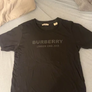 Svart Burberry t-shirt med tryck - Svart t-shirt från Burberry med klassiskt tryck 'BURBERRY LONDON ENGLAND' på bröstet. T-shirten har rund halsringning och korta ärmar. Tillverkad i mjuk bomull för en skön känsla och stilren look.