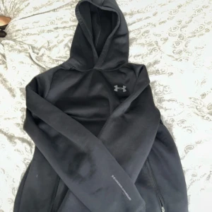 Svart hoodie från Under Armour YXL - Svart hoodie från Under Armour med klassisk logga på bröstet. Tröjan har huva, långa ärmar och dragkedjefickor. Tillverkad i mjukt och värmande fleece-material, perfekt för kyliga dagar eller när du vill ha en sportig look.