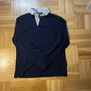 Mörkblå pikétröja från Polo Ralph Lauren - Klassisk mörkblå långärmad pikétröja från Polo Ralph Lauren med vit krage och knäppning framtill. Tröjan har det ikoniska röda broderade logotypen på bröstet och är tillverkad i mjuk bomull. Perfekt för en stilren och avslappnad look.