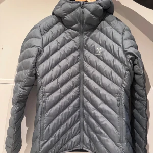 Grå dunjacka från Haglöfs - Snygg grå dunjacka från Haglöfs med chevronmönstrad quiltning och hög krage. Jackan har två dragkedjeförsedda fickor och hel dragkedja framtill. Perfekt för kyliga dagar och riktigt skön att bära.Nypris 2300kr 