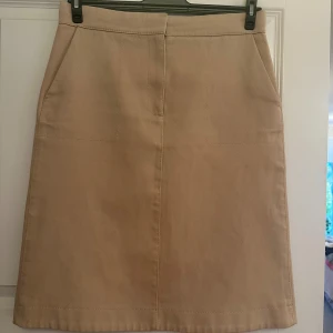 Beige A-linje kjol från COS - Stilren beige A-linjeformad kjol från COS i 100% bomull. Kjolen har två stora fickor framtill och en klassisk midja. Perfekt för dig som gillar minimalistisk och clean stil.