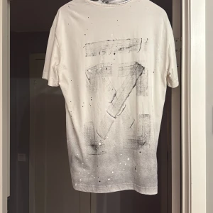 Off-White vit t-shirt med tryck - Vit t-shirt från Off-White med svart spray paint-effekt och stänkdetaljer. Stor Off-White-logga på ryggen och mindre tryck framtill. Klassisk passform och rund halsringning. Perfekt för dig som gillar streetwear och unika prints.