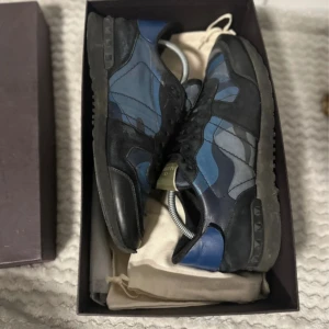 Valentino Rockrunner sneakers blå/svart - Säljer ett par Valentino Rockrunner sneakers med blå och svarta camouflage-paneler, svarta mockadetaljer och platt sula. Skorna har snörning och ikoniska nitar på hälen. Materialmix av skinn och mocka ger en lyxig känsla. Perfekt för dig som gillar statement-sneakers. Storlek 43, Små defekter, skriv för bilder. Inget og 