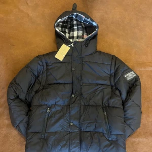 Svart pufferjacka från Burberry - Svart pufferjacka från Burberry med huva och rutig innermössa i svart, vitt och rött. Jackan har dragkedjor på fickorna, tryckknappar framtill och en stor Burberry London England-patch på ärmen. Perfekt för kalla dagar och riktigt snygg streetstil.