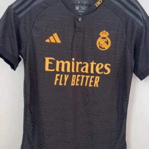 Svart Real Madrid fotbollströja Adidas - Snygg svart Real Madrid fotbollströja från Adidas med gul logga och tryck. Tröjan har korta ärmar, subtilt mönster i tyget och klassiska Adidas-ränder på axlarna. Materialet är lätt och andas, perfekt för träning eller match.