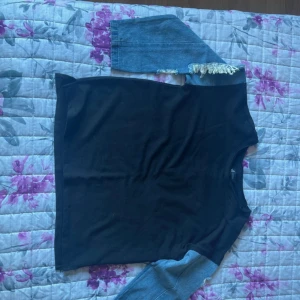 Svart tröja med jeansärmar - Cool svart tröja med kontrasterande jeansärmar och fransdetaljer vid axlarna. Tröjan har en rak passform och rund halsringning, perfekt för dig som vill sticka ut med en unik stil.