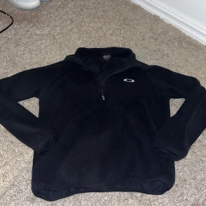 Svart fleece-tröja från Oakley XS - Svart Oakley fleece-tröja i storlek XS med halv dragkedja och diskret logga på bröstet. Perfekt för kyliga dagar, med lång ärm och mjukt material som håller dig varm. Enkel och stilren design som funkar till det mesta.