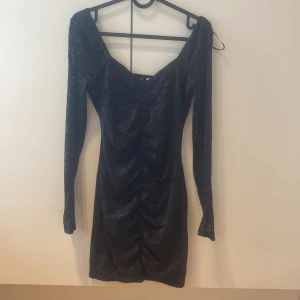 Svart spetsklänning från Divided - Supersnygg svart kort klänning från Divided med lång ärm och v-ringad hals. Klänningen har spetsdetaljer och rynk framtill som ger en riktigt schysst siluett. Perfekt för dig som vill sticka ut lite extra med en elegant men edgy look.