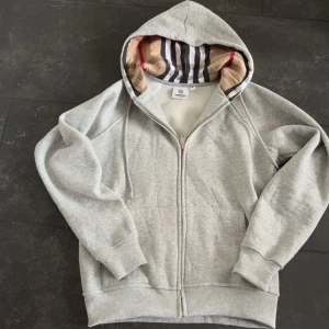 Grå hoodie från Burbery med dragkedja - Snygg grå hoodie från Burberry med klassisk rutmönstrad huva och broderad logga på bröstet. Helt ny burrberry hoodie (äkta) inte för ett jättebra pris HELT NY